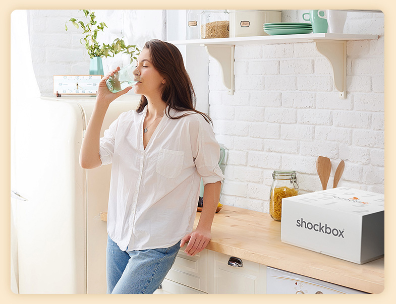 Donna in cucina che beve il drenante da un bicchiere di acqua con affianco Shockbox